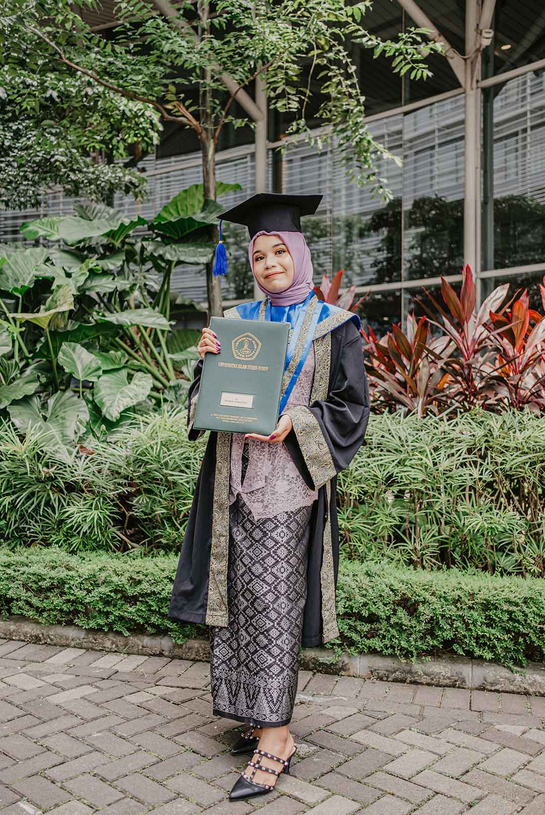 Foto wisuda perseorangan dengan toga dan map ijazah di taman kampus